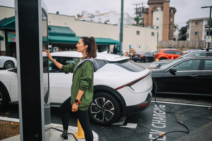 Woman stops at the EV charging station, checking the display screen while the car is parked and recharged, on a parking lot in front of the strip mall.